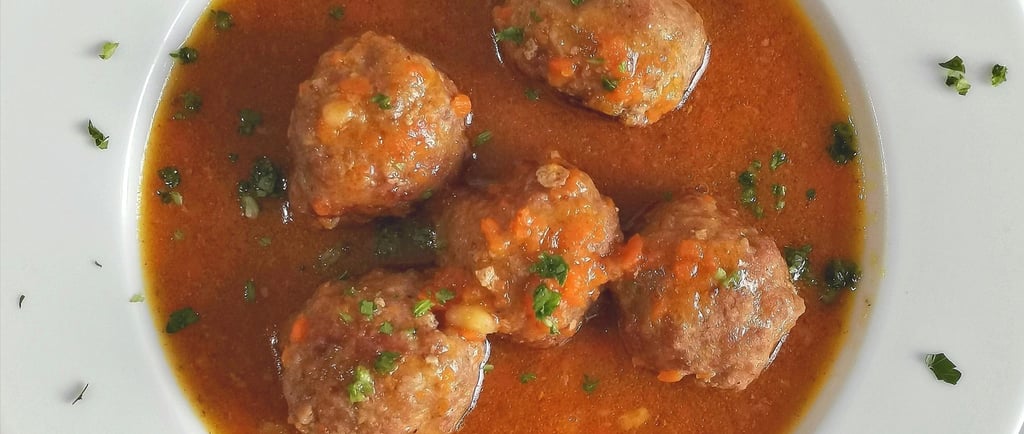 Beef meatballs in savory tomato vegetable broth served in a white bowl with herbs.
