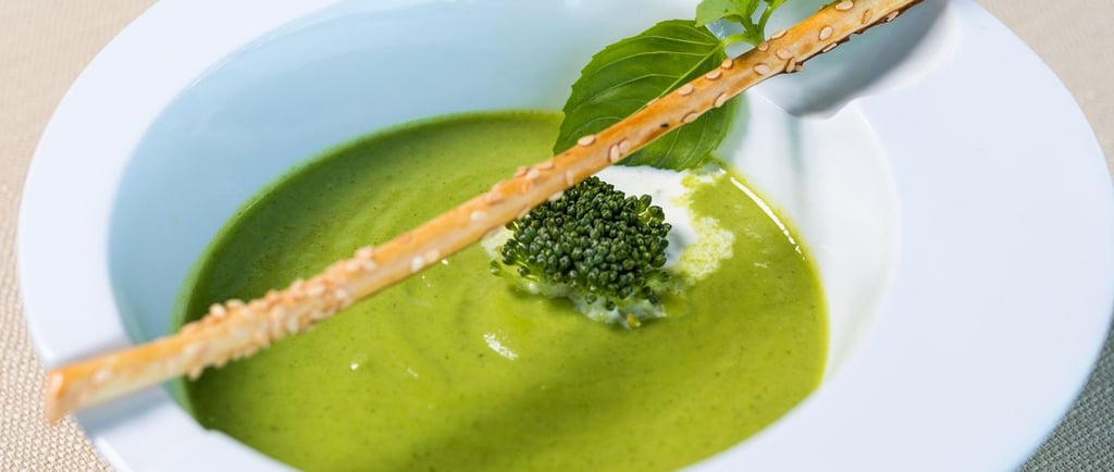 Creamy green broccoli soup in a white bowl garnished with a breadstick and fresh herbs.