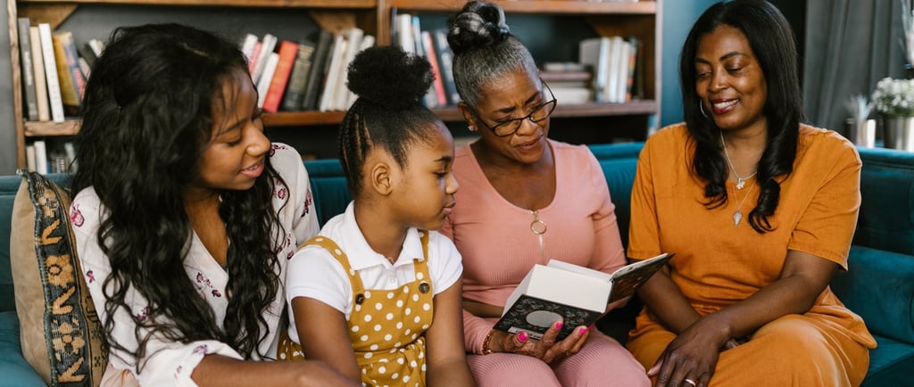 Three generations of Black women sitting on a sofa reading a book together in a library.