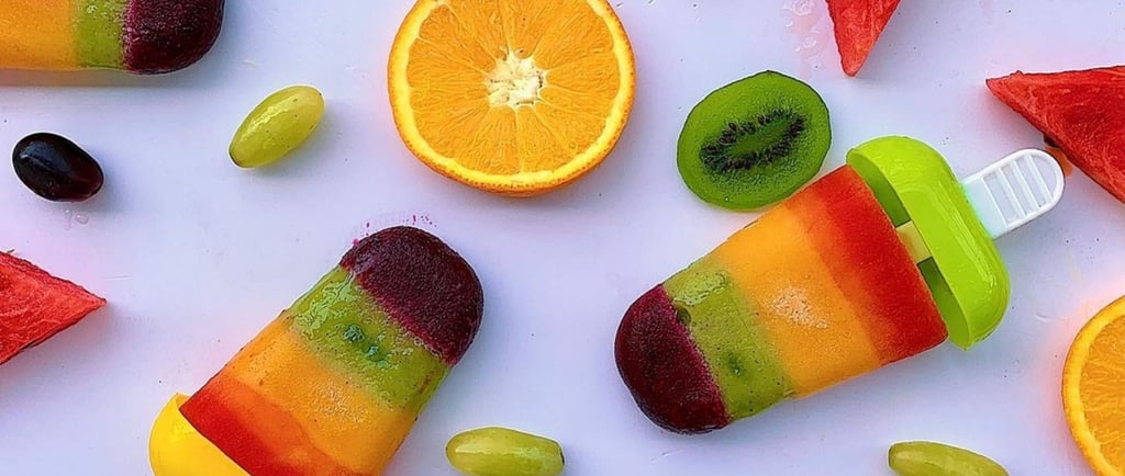 Layered fruit juice popsicles surrounded by fresh orange, kiwi, and watermelon slices on a white surface.