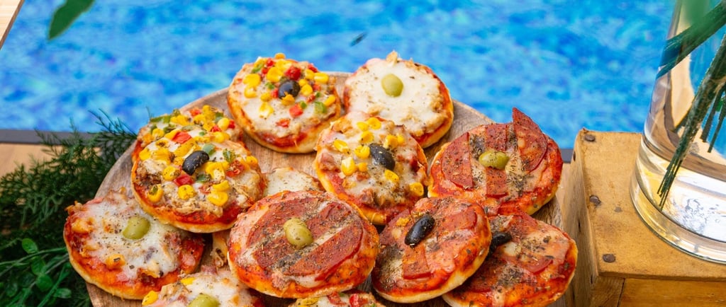 Mini pizzas with assorted toppings on a wooden platter by a blue swimming pool.