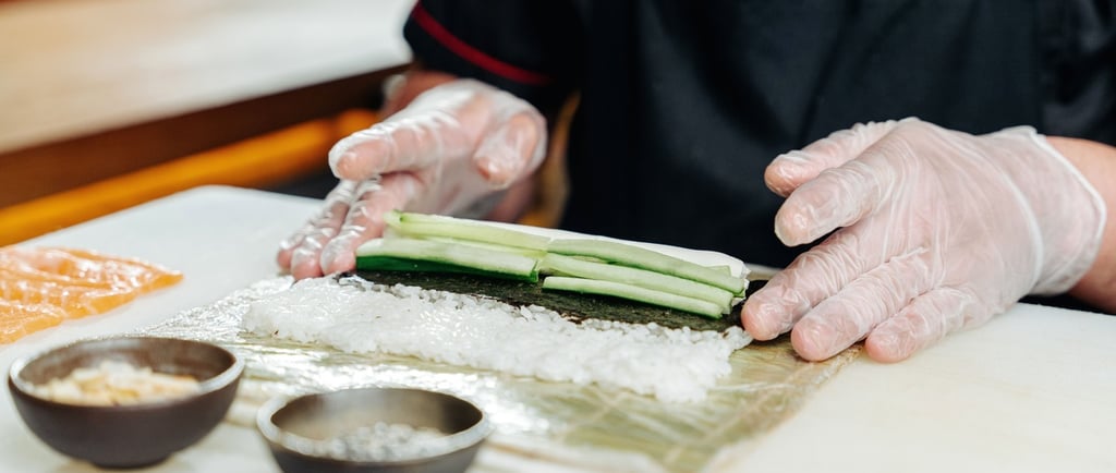 Professional chef wearing gloves preparing a fresh sushi roll with cucumber, rice, and nori seaweed.