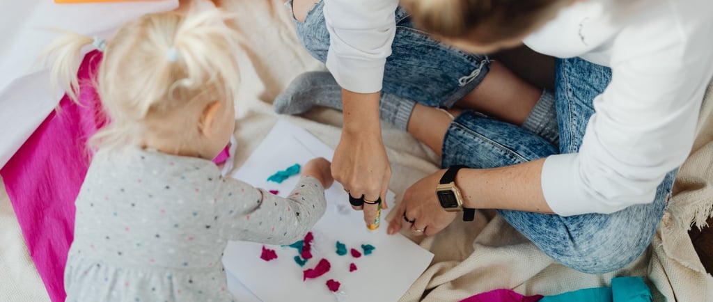Mother and toddler girl creating colorful paper crafts with glue and tissue paper on a blanket.