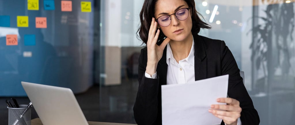 Stressed businesswoman in glasses reviewing financial documents at her office desk with a laptop.