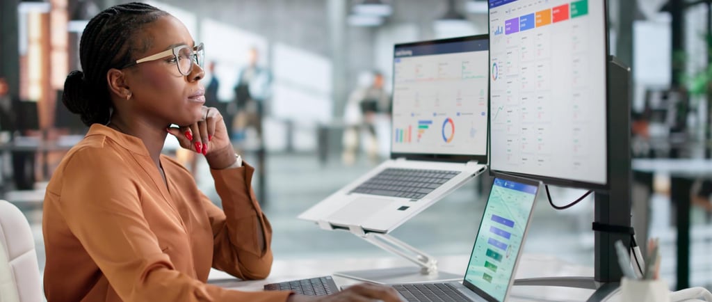 A female data analyst reviews a CRM system dashboard on multiple monitors in a modern office.