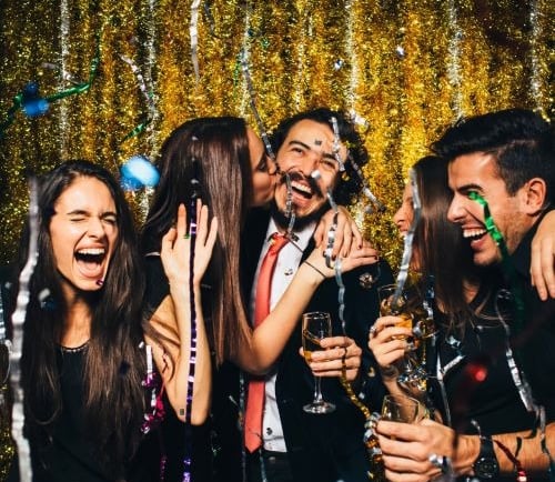 Cheerful friends celebrating at a New Year's Eve party with confetti and gold tinsel background.
