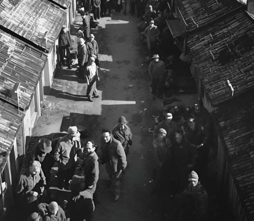 Historical black and white aerial view of a narrow alley between wooden barracks with people standing.
