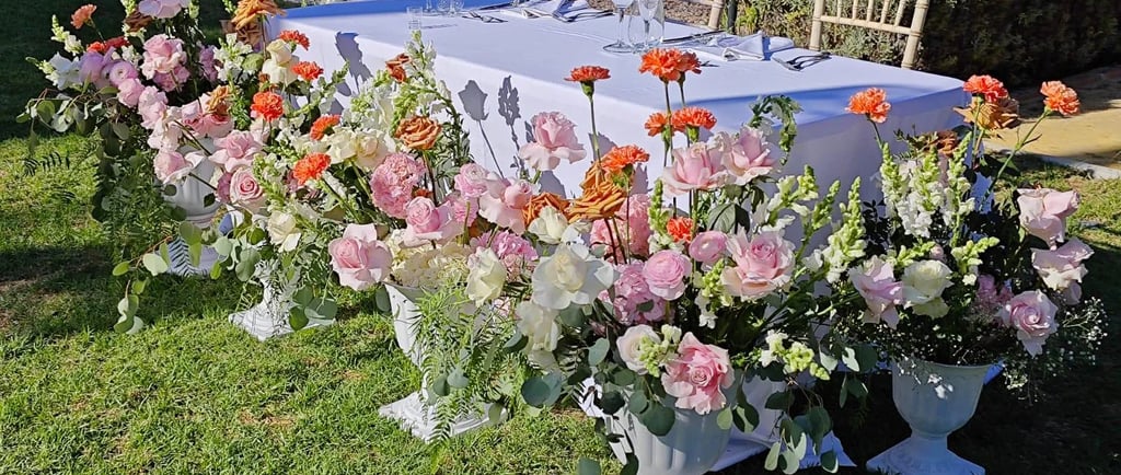 An outdoor wedding table elegantly decorated with lush pink and orange floral arrangements in white urns on a grassy lawn.