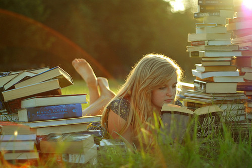 Image of a blonde girl surrounded by books reading a book in the middle of an open field