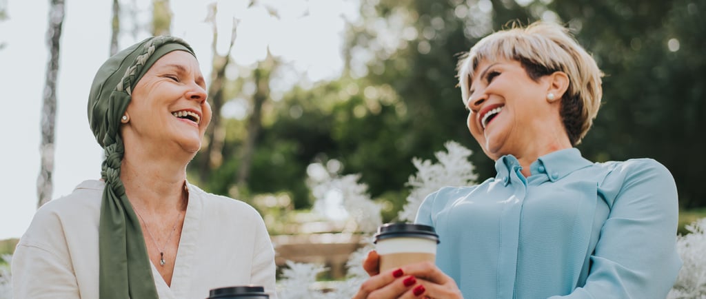 two women sitting on a bench in a park laughing and drinking coffee