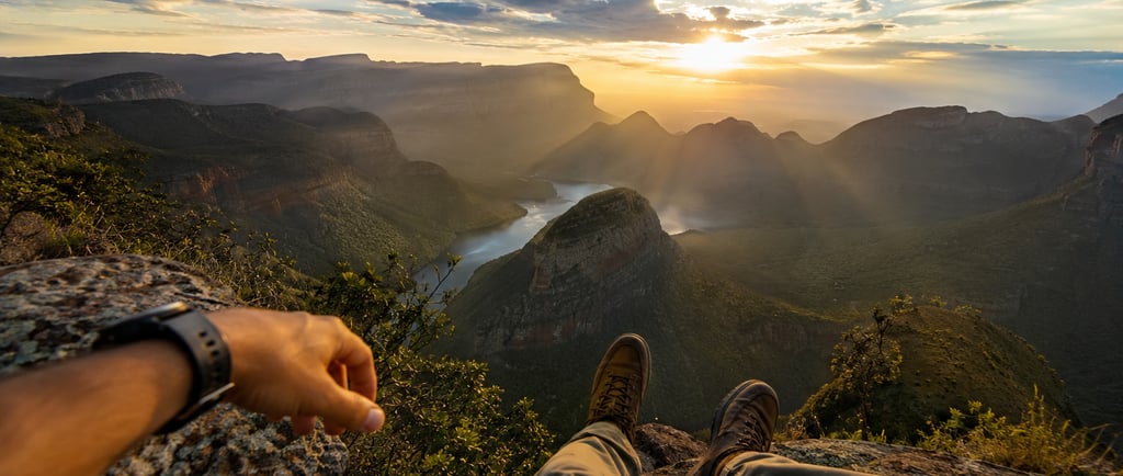 Hiker sitting on a rocky cliff edge overlooking a scenic mountain valley and river at sunrise.