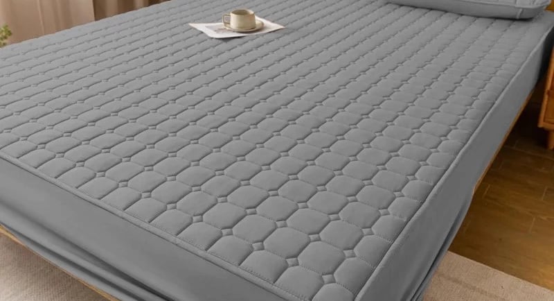Quilted grey waterproof mattress protector with matching pillow shams on a modern wooden bed frame.