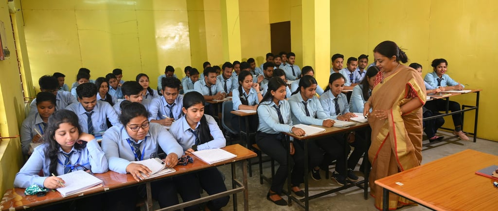Students studying in a classroom with a lady teacher guiding them.