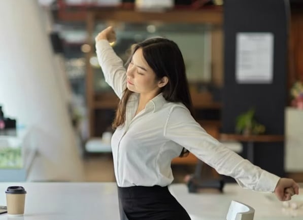 Business woman in white shirt stretching at her office desk to relieve stress and back pain.