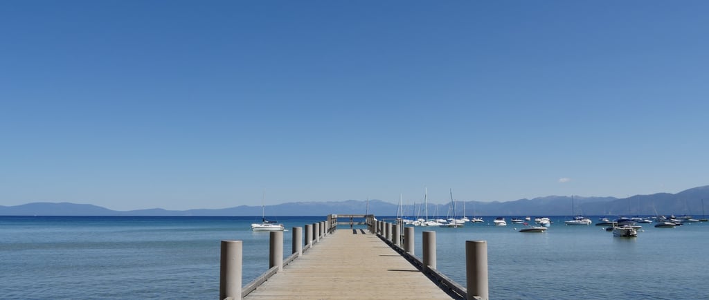 A dock with wooden bridge leading to a water body and ships docked in distant background.