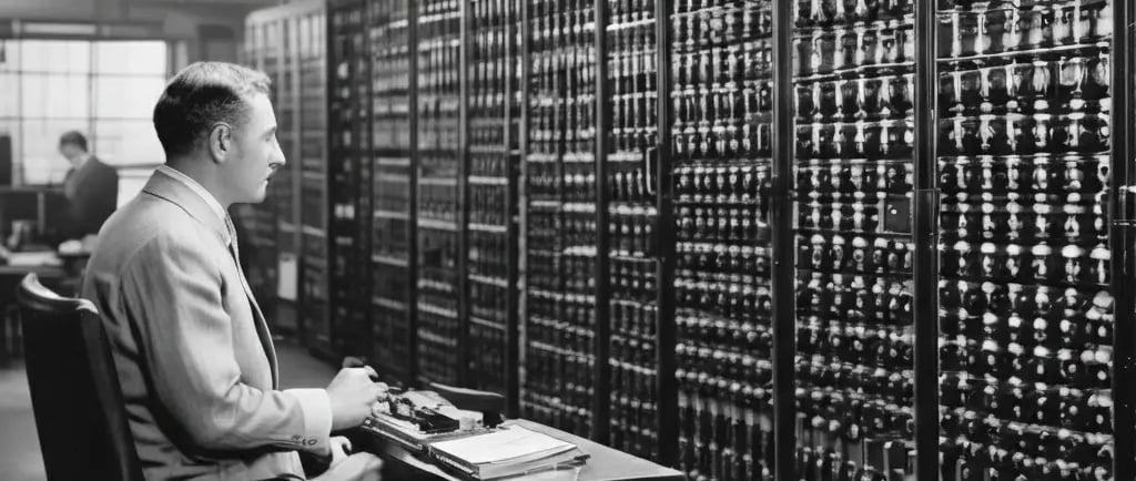 Homem operando o ENIAC em laboratório da década de 1940, sentado diante de grandes painéis repletos