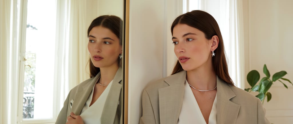 Elegante Frau mit minimalistischem Silberschmuck – Kette, Ohrringe, Armband – steht vor Spiegel im modernen Raum