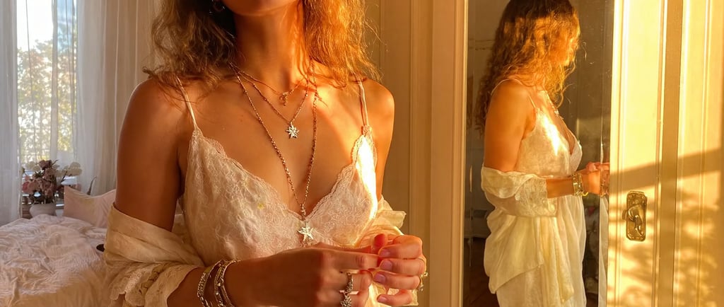 Blonde boho-style model with copper-orange hair standing in front of a vintage mirror in a sunlit bedroom. She wears a flowin