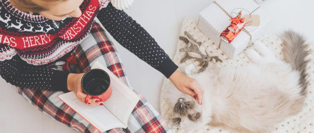 Moteris su kalėdine pižama ir megztiniu sėdi ant grindų, glosto katę, rankoje puodelis.