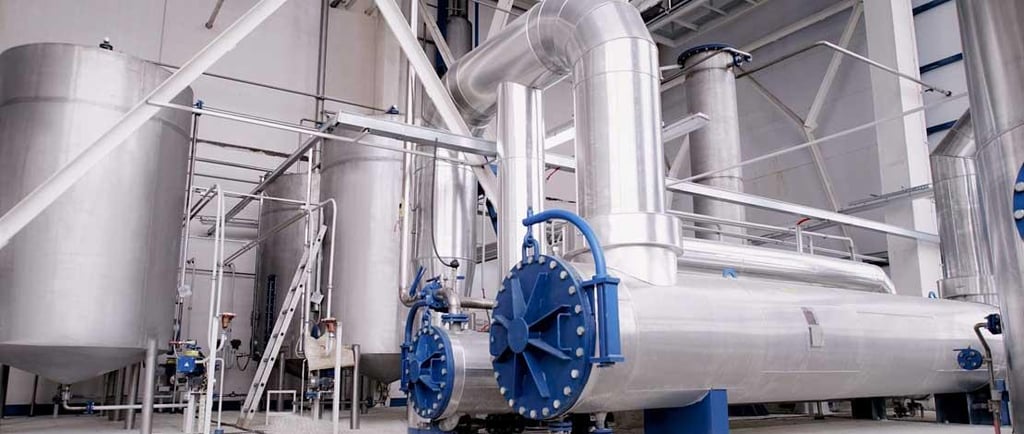 Industrial factory interior featuring stainless steel storage tanks and large insulated piping systems.
