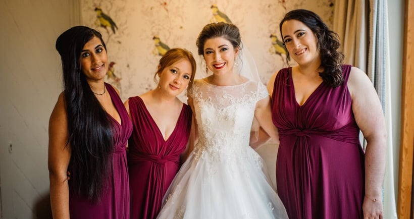 Smiling bride in a white lace wedding gown posing with bridesmaids in plum-colored dresses.