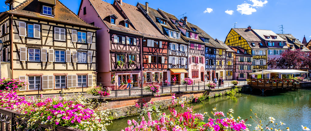 Historic colorful half-timbered houses line a tranquil canal in Colmar, France, adorned with vibrant flowers on a sunny day.