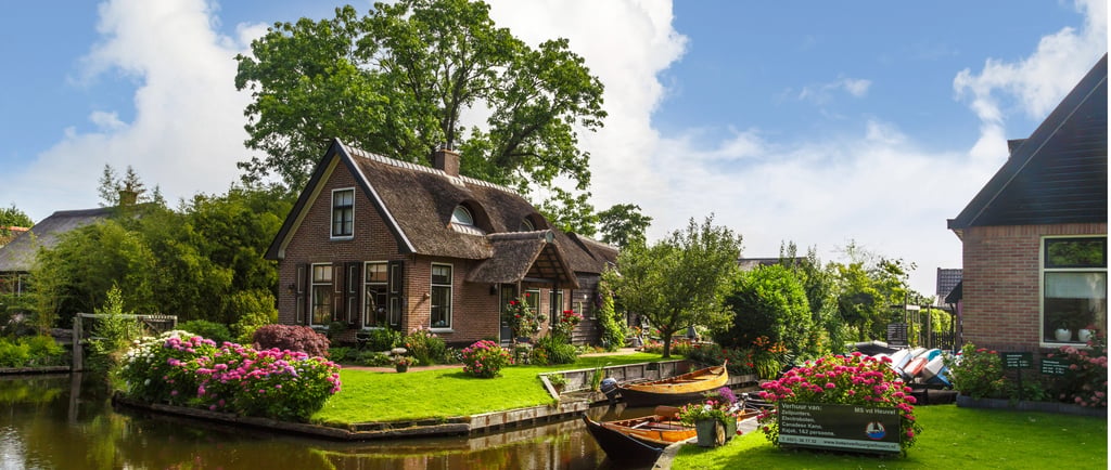 Quaint thatched roof cottage in Giethoorn, Netherlands, with wooden boats on a canal and blooming pink flowers.