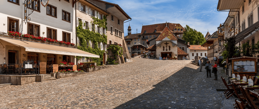 Charming cobblestone street in Gruyères Switzerland with historic buildings and outdoor cafe seating under a blue sky.
