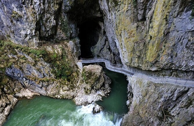 A metal walkway bridge leads into the dark mouth of the Skocjan Caves over a flowing green river.