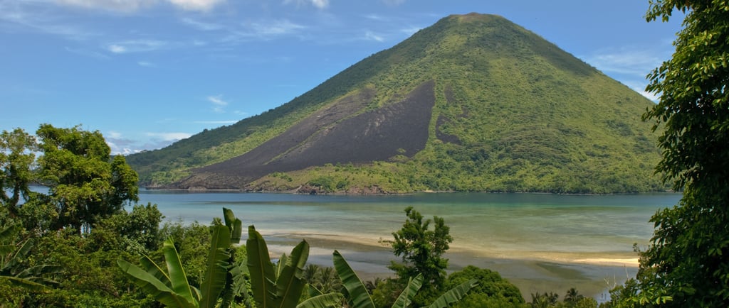 A lush tropical volcano with dark lava flows overlooks a turquoise ocean bay under a blue sky.