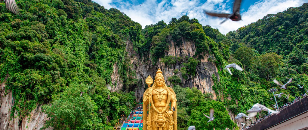 The gold Lord Murugan statue at Batu Caves in Malaysia with limestone hills and pigeons in the foreground.