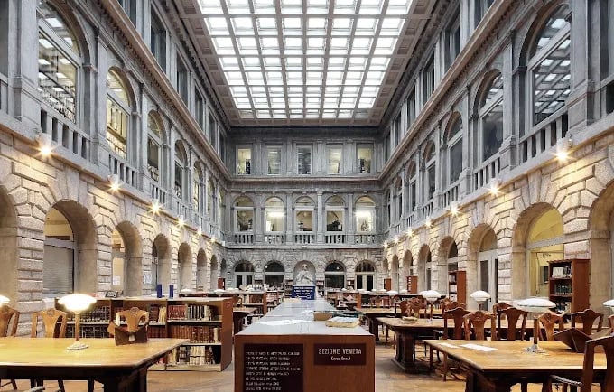 Historic Italian library interior with classic wooden study desks and a large glass atrium ceiling.