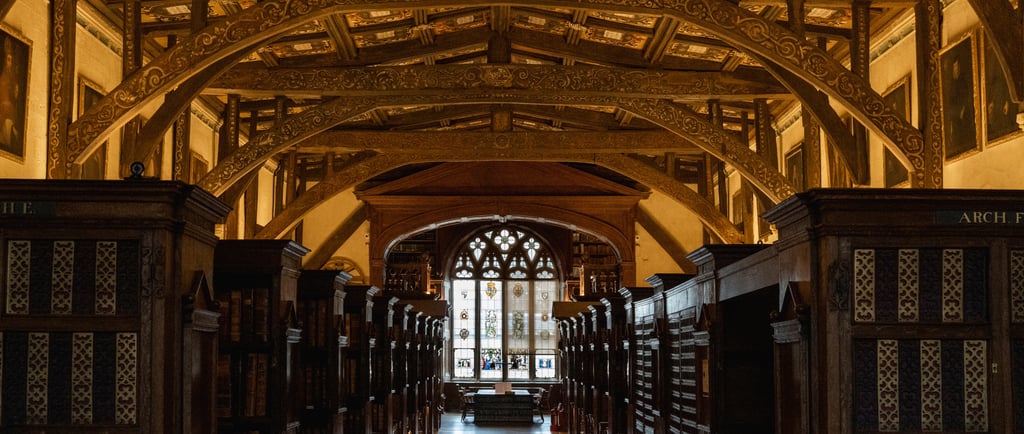 Ornate wooden arched ceiling over rows of bookshelves in a historic library with stained glass windows.