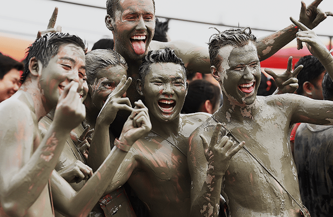 A group of friends covered in mud laughing and gesturing at a mud festival or obstacle course race.
