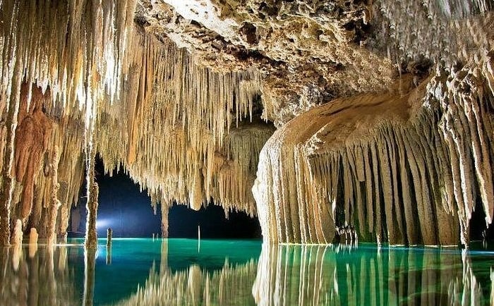 Underground limestone cavern featuring sharp stalactites reflecting in a clear turquoise cenote pool.