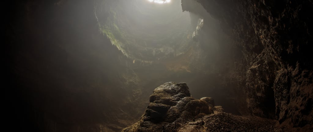 Sunlight streams through a hole in the ceiling of a dark limestone cave onto a central rock formation.
