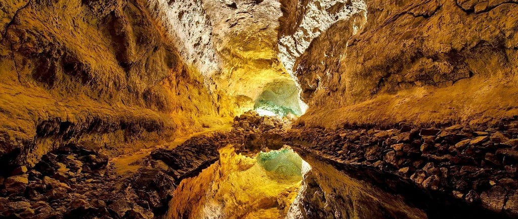 Inside an ancient volcanic cave with a reflective pool on the floor and warm light illuminating the rock formations.