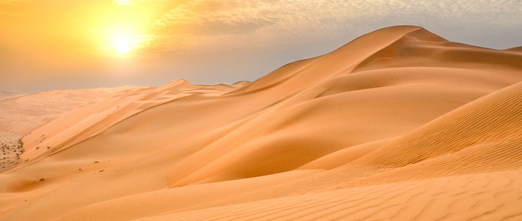 Golden sunset over rolling sand dunes in a vast desert landscape with a glowing sun and hazy sky.