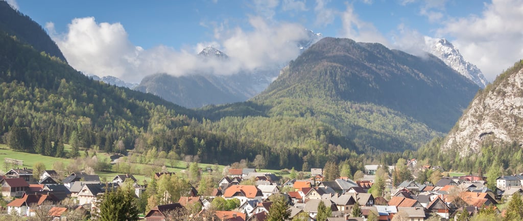 Panoramic view of a scenic mountain village in the Julian Alps with snow-capped peaks and pine forests.