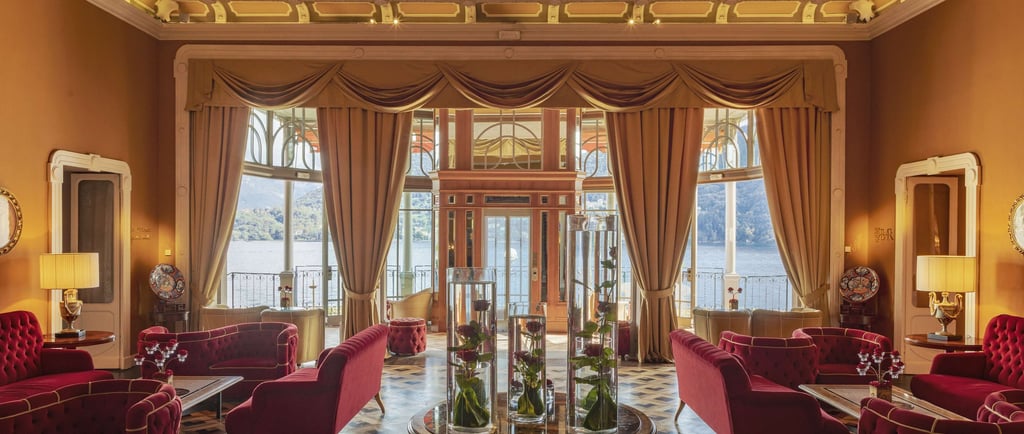 Elegant hotel lounge with red velvet sofas and a panoramic view of Lake Como through large windows.
