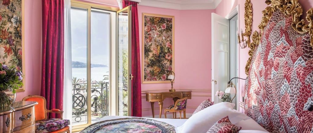 Luxury pink hotel room with crystal chandelier, ornate gold headboard, and balcony overlooking the sea.