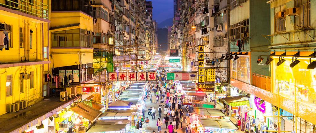Crowded night market in Hong Kong with glowing neon signs and street food stalls between high-rise buildings.