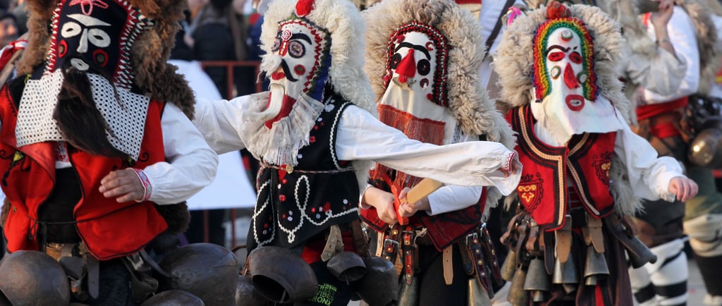 Performers in traditional Bulgarian Kukeri costumes and masks dancing with large bells during a festival.