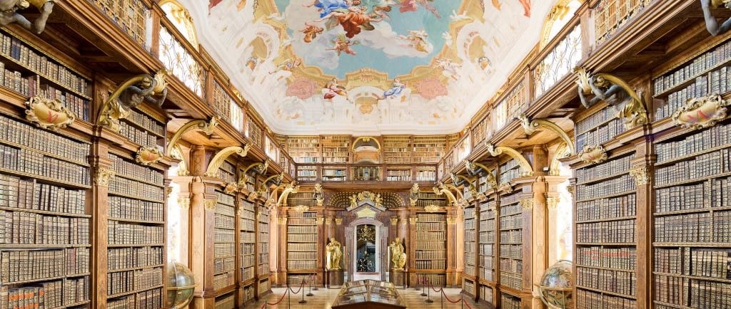 Ornate Baroque library with wooden bookshelves and a colorful fresco painted on the ceiling.