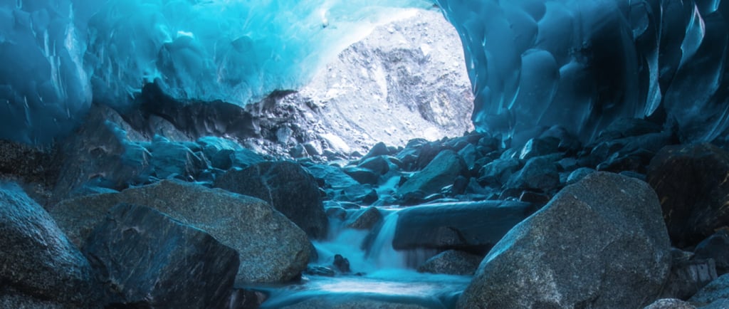 A flowing stream through a glowing blue glacial ice cave with large rocks and a mountain view.