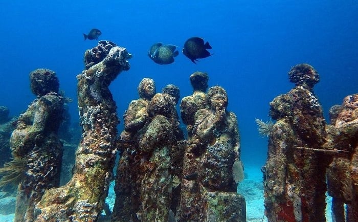 Underwater statues covered in coral reef at an ocean sculpture park with tropical fish swimming nearby.