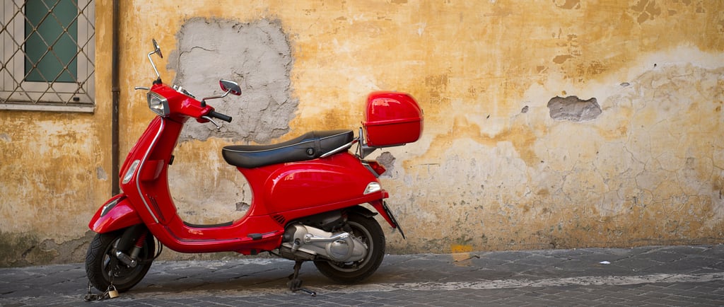 A classic red Italian motor scooter parked against a rustic, yellow textured wall on a cobblestone street.