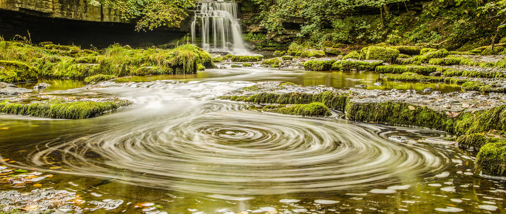 Long exposure of a swirling water vortex in a river below a scenic forest waterfall.