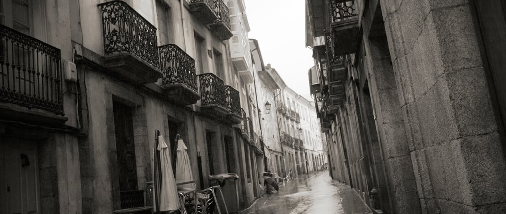 A narrow cobblestone European street in black and white with ornate iron balconies and wet pavement.