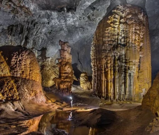 A cavern explorer with a headlamp stands amidst massive limestone stalagmites and rock formations inside a dark cave.